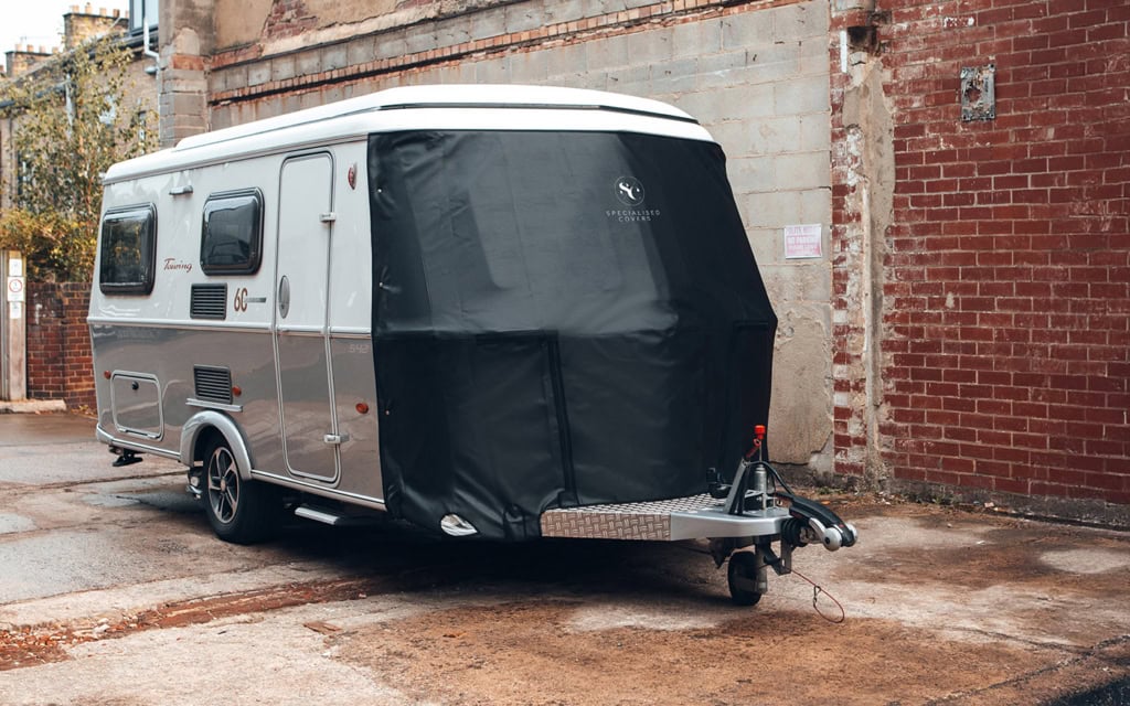Specialised Covers fitted on a caravan for protection, with a black cover over the front and side windows, parked on a concrete surface near a brick wall.
