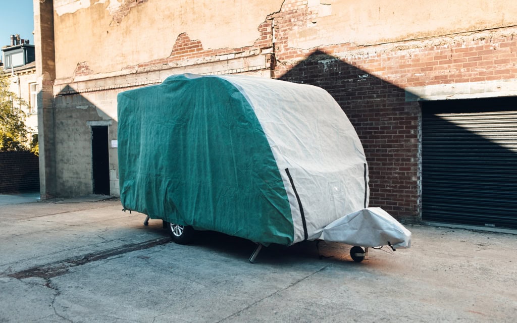 Caravan covered with a protective green and grey cover parked on a street near a brick building.