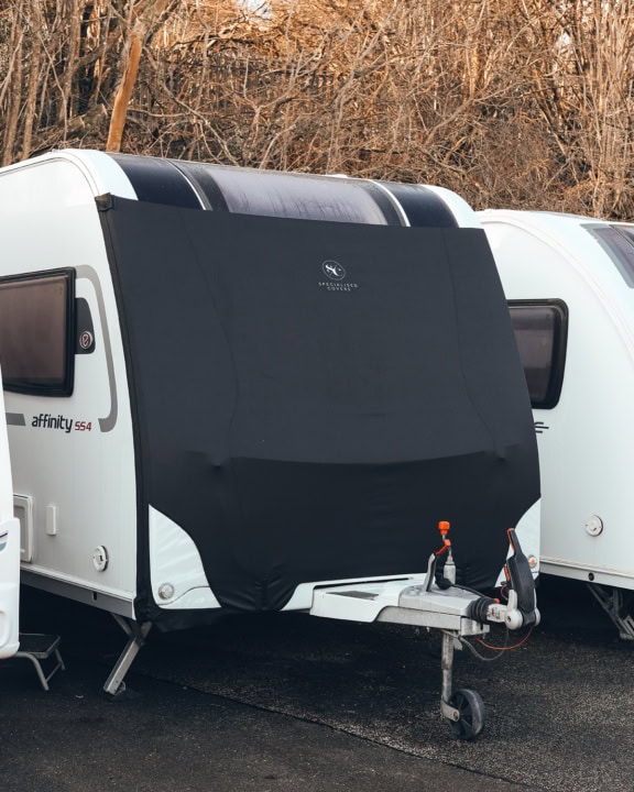 Black caravan cover fitted over a white caravan, protecting it from weather elements, with a hitch and towing equipment visible in the foreground.