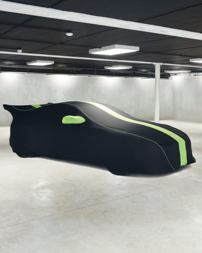 A picture of an Aston Martin in a garage with a Black Indoor Plus cover on with green stripes, piping and wing mirrors.