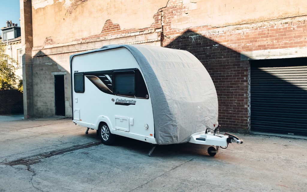 Caravan with a grey protective cover parked on a street near a brick building.