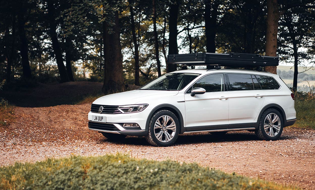 Enhanced fit Volkswagen estate car with custom outdoor protective cover on gravel path in woodland setting.
