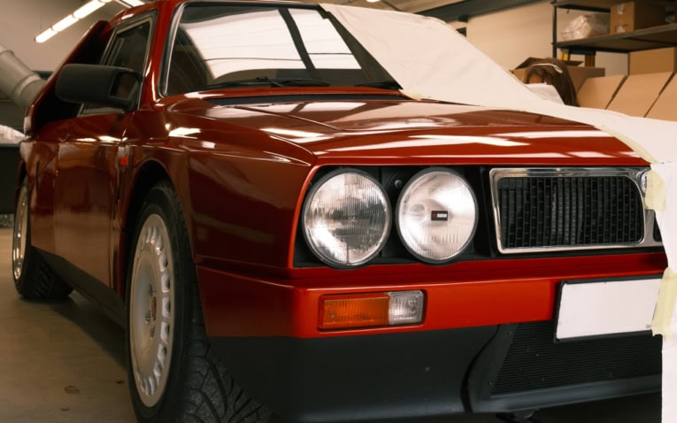 A classic red Lancia Montecarlo in a workshop, covered partially with protective masking for maintenance or restoration, highlighting specialised vehicle covers and automotive care.