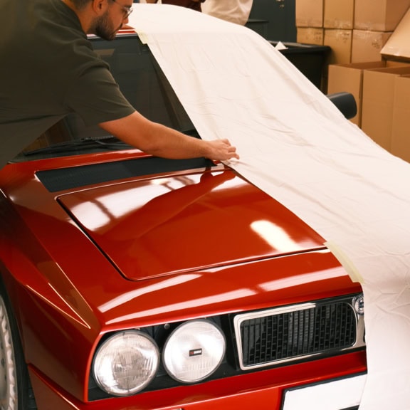 Durable custom car cover being fitted to a classic red vehicle, providing weather protection and maintaining interior and exterior condition. Perfect for classic car enthusiasts and dealerships.