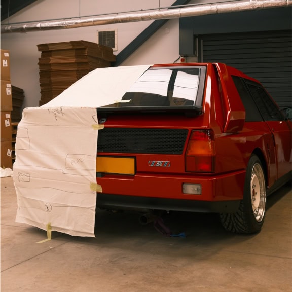 1. Custom car cover being prepared for a classic red rally car in a workshop, with protective material partially covering the vehicle.