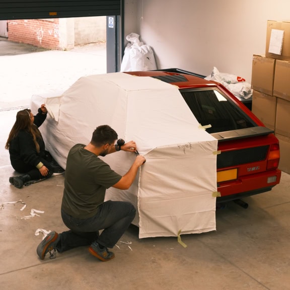 Protective car cover being fitted to sports car in a garage for optimal vehicle protection and storage.