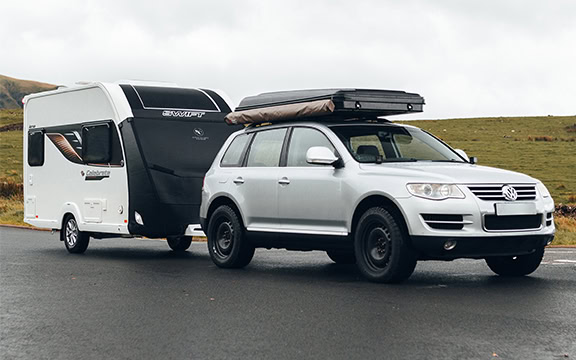 Silver SUV towing a caravan with a protective cover, equipped with roof storage, parked on a scenic roadside. Perfect for protecting your caravan during travel.