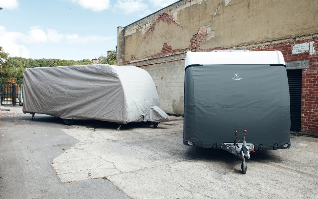 Two caravan covers protecting caravans parked outdoors near a brick wall and trees.