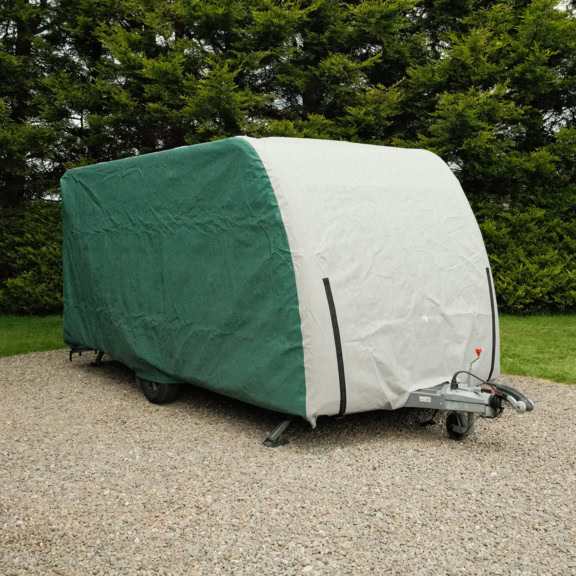 Green and grey caravan cover protecting a caravan outdoors on a gravel surface with trees in the background.