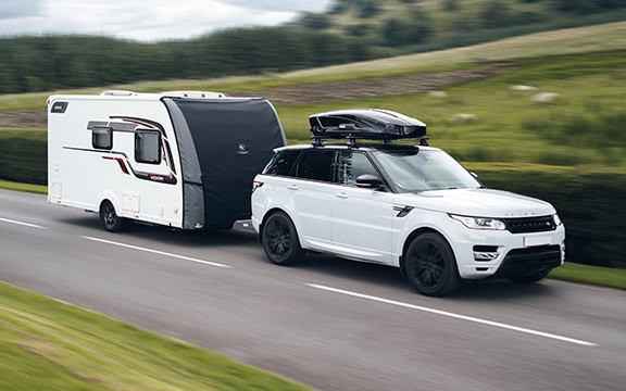 White SUV towing a caravan with a tailored cover on the caravan, driving on a scenic road with green fields and trees in the background.