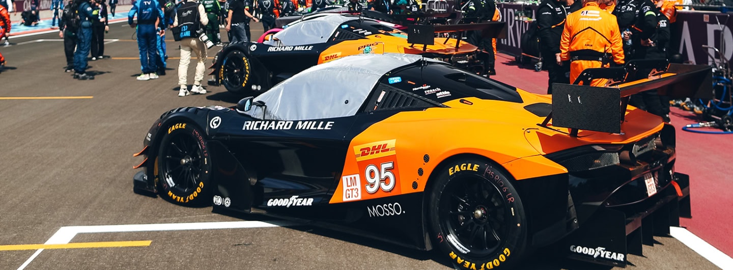 Two high-performance race cars parked in the pit lane at a racing event, with team members and officials nearby.