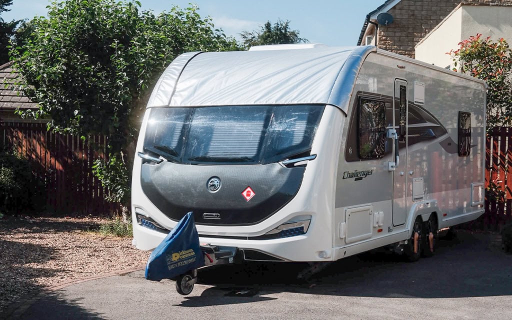 Modern caravan with panoramic window cover parked on a driveway with trees and houses in the background.