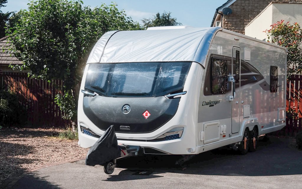 Modern caravan with panoramic window cover parked on a driveway with greenery in the background.